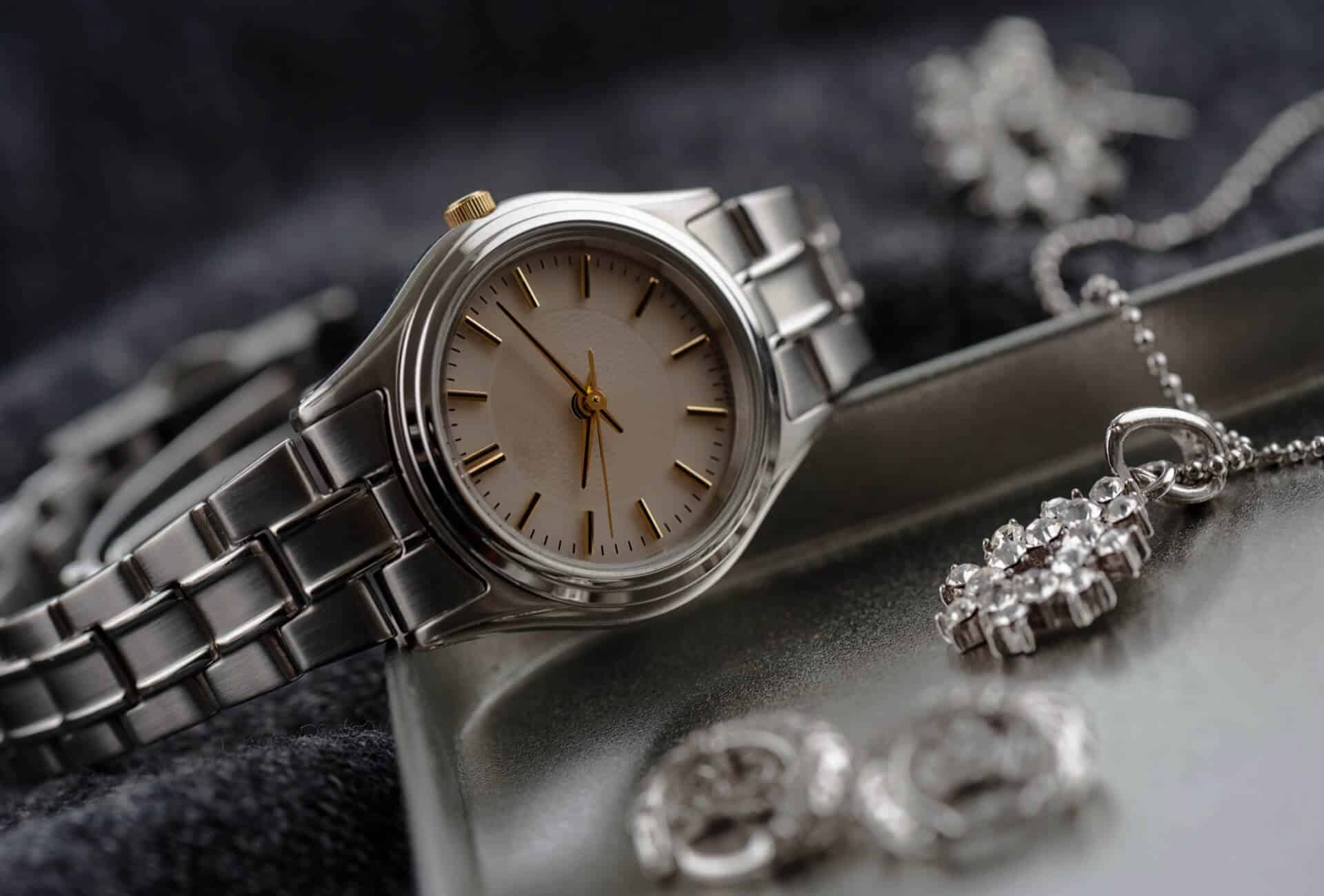  A silver wristwatch with a champagne-colored face sits on a metal tray next to a silver necklace and earrings.